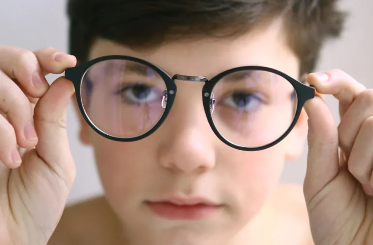 Close-up of a boy holding eyeglasses in front of his face, with his eyes visible through the lenses, representing vision correction or eye care for children.