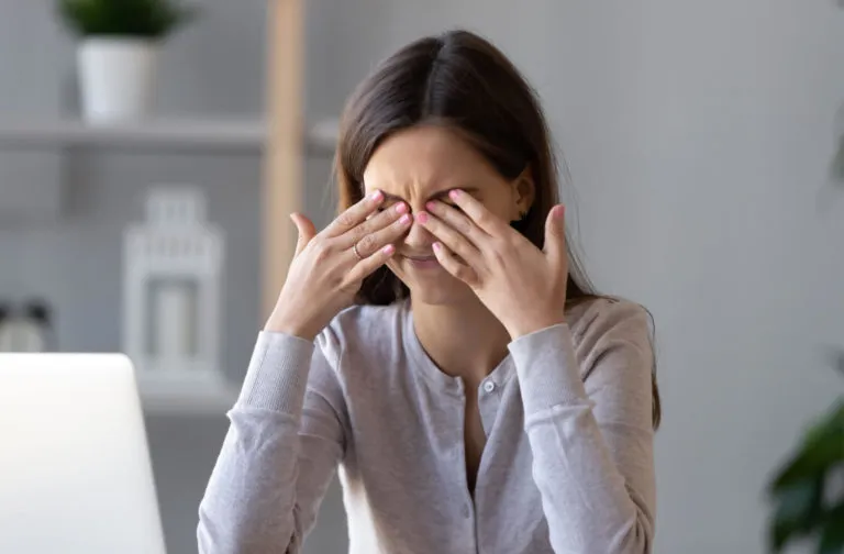 A young woman rubbing her eyes with both hands while sitting at a desk.