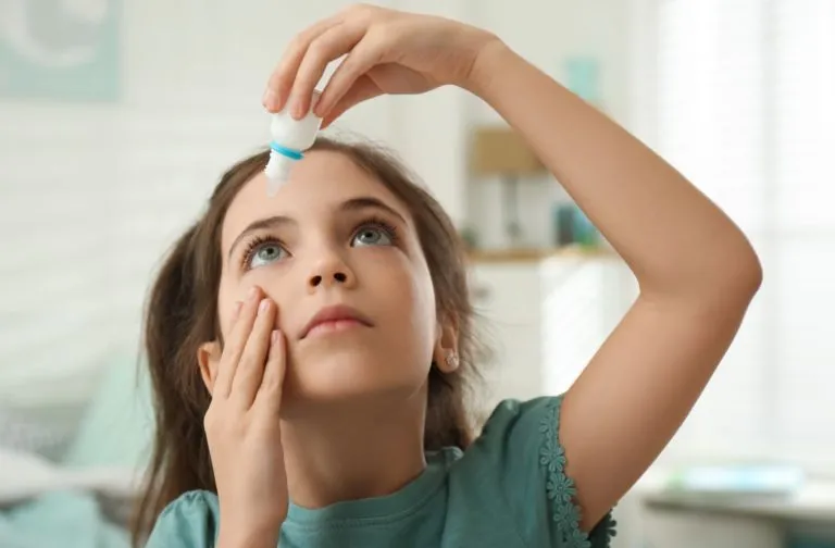Young girl using eye drops at home, holding the bottle above her eye and looking up, representing eye care or dry eye relief in children.