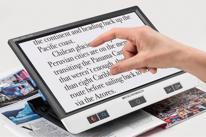 Hand using an Eschenbach electronic magnifier to enlarge printed text on a magazine, demonstrating a digital low vision aid for easier reading.