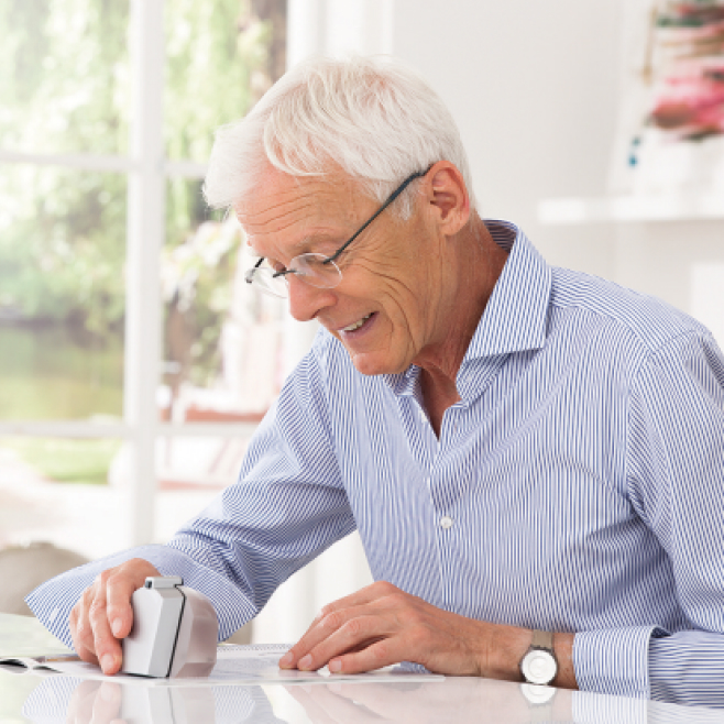 Older man using a portable low vision aid device to read printed text at home, demonstrating assistive technology for visual impairment.