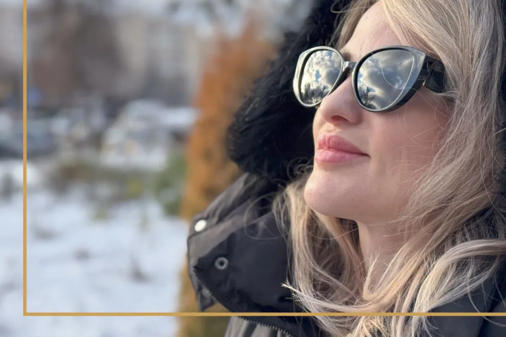 A woman looks at the sun during the winter while wearing sunglasses.
